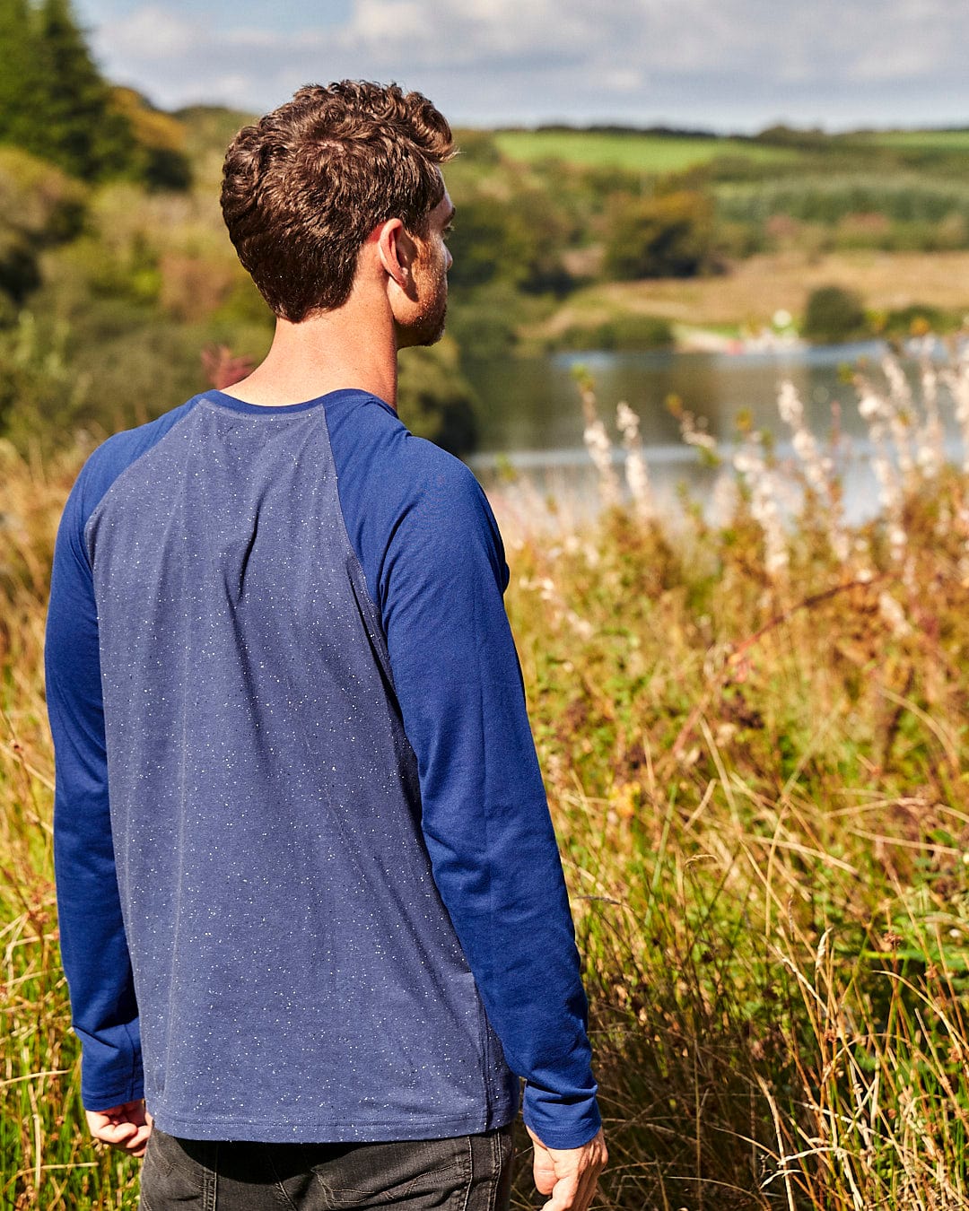 A man standing in a field looking at a lake, wearing a Saltrock Home Run Nep - Mens Raglan Long Sleeve T-Shirt - Blue.