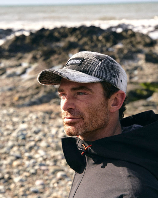 A man wearing a Saltrock Hunter - 5 Panel Cap - Grey standing on a rocky beach.