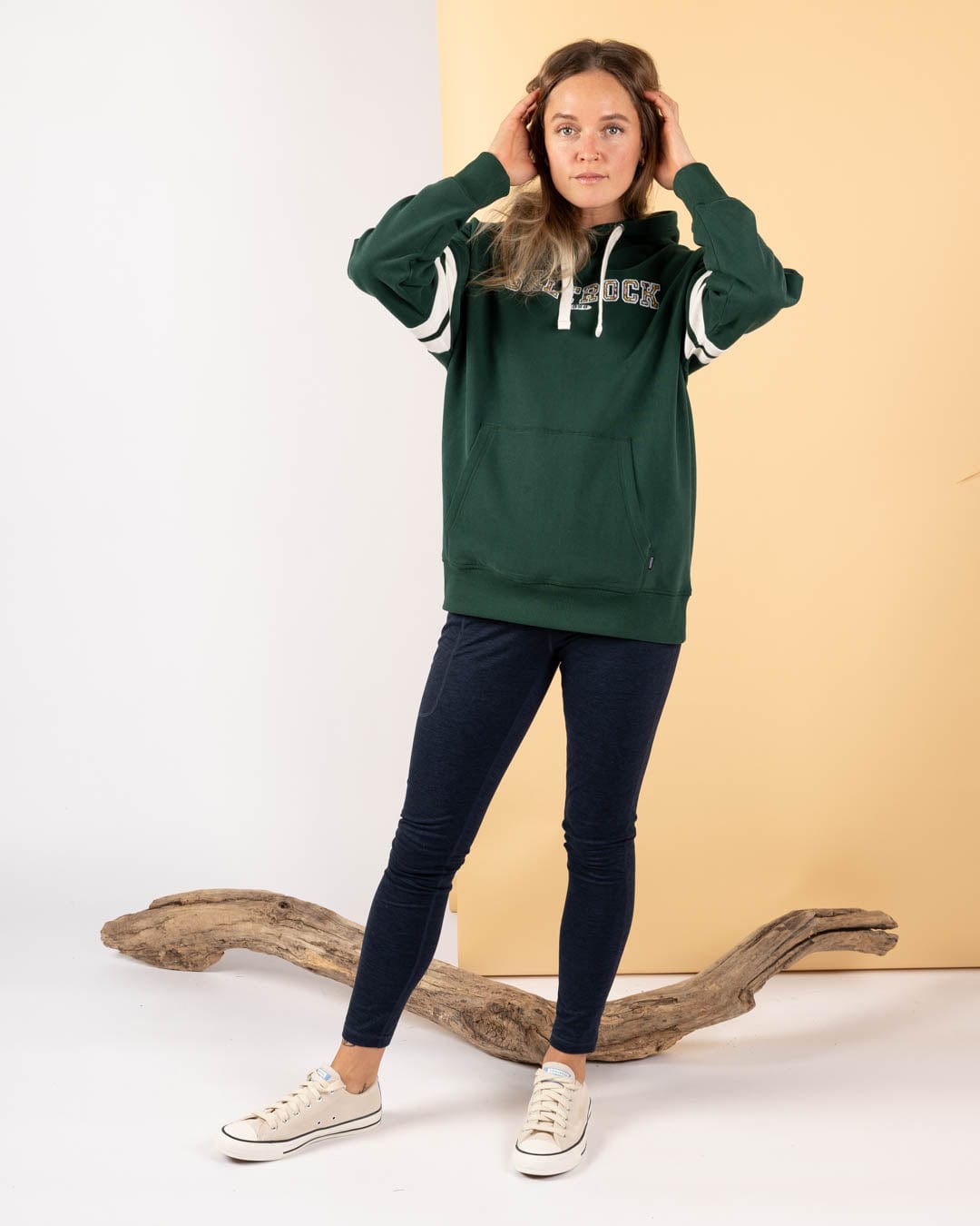 A woman stands indoors wearing the Saltrock Campus Floral Women's Pop Hoodie in dark green, styled with dark leggings and white sneakers. Driftwood rests on the floor behind her against a pale yellow background.