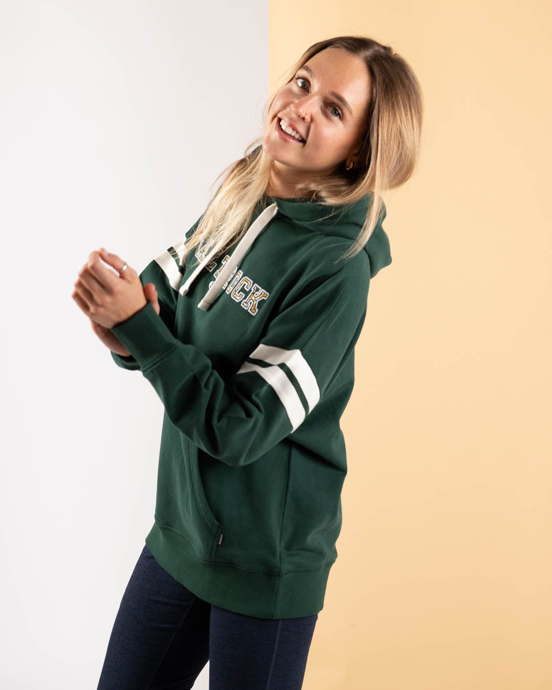 A woman wears the Saltrock Campus Floral Women's Pop Hoodie in dark green, featuring white stripes and lettering, while standing against a split white and beige background, smiling at the camera.