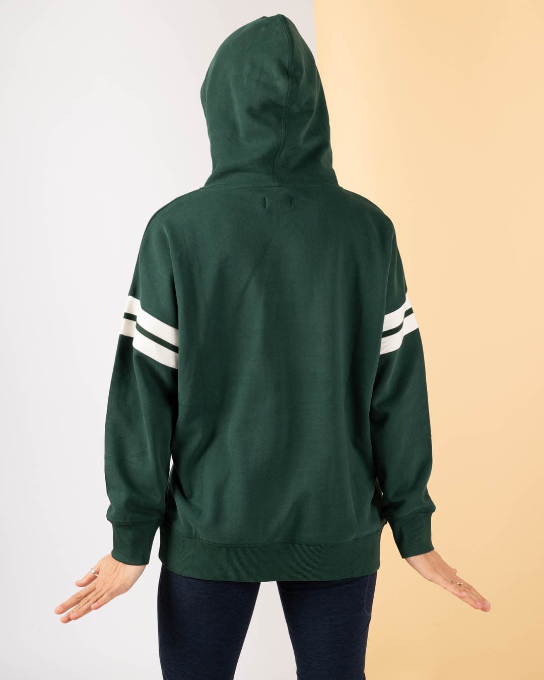 A person wearing the Saltrock Campus Floral - Womens Pop Hoodie in dark green, made from 100% cotton with white sleeve stripes, stands with their back to the camera against a white and beige background.
