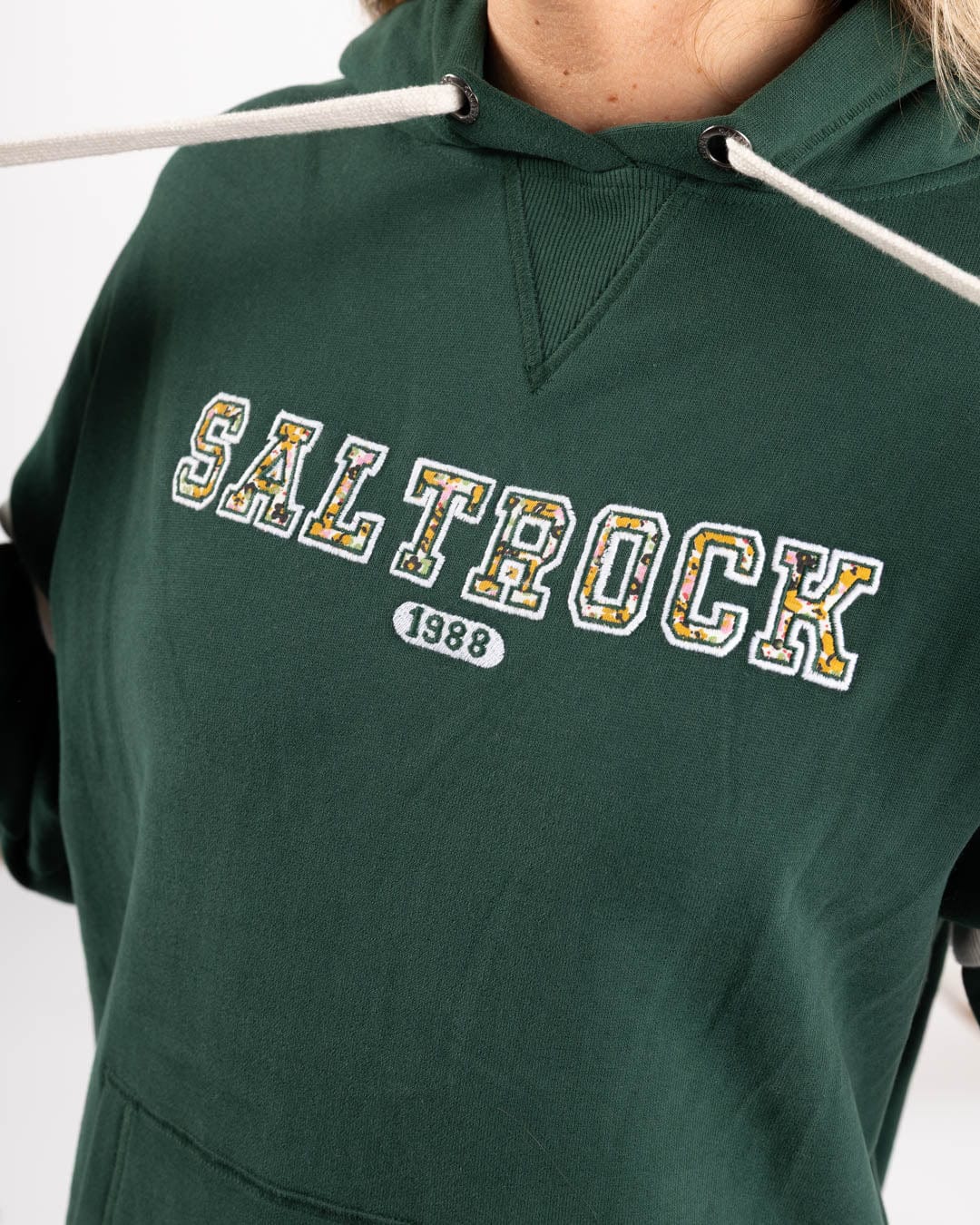 A woman wears the Saltrock Campus Floral Women’s Pop Hoodie in dark green, made from 100% cotton, featuring "SALTROCK" and "1988" printed on the front with white and yellow outlined letters.