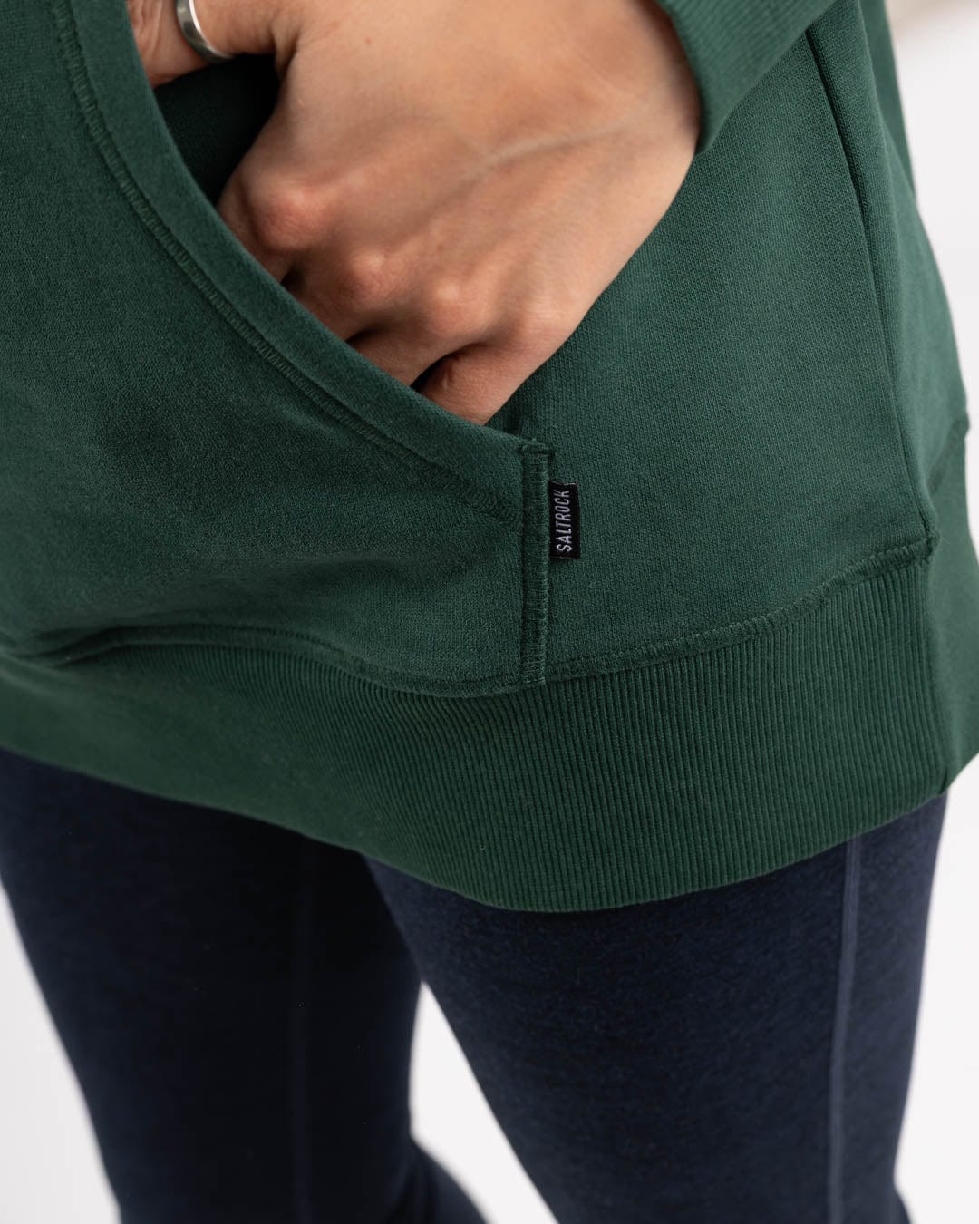 A person wears the Saltrock Campus Floral Women's Pop Hoodie in dark green, made from 100% cotton, with one hand in the pocket, paired with dark blue pants or leggings.