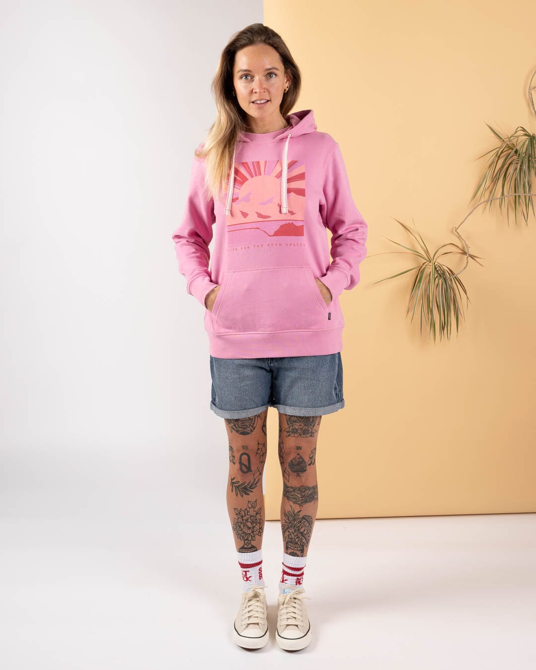 A woman faces the camera wearing the Saltrock Open Spaces - Womens Pop Hoodie in pink, styled with denim shorts and sneakers. She showcases leg tattoos against a two-tone backdrop with plants, capturing a laid-back Saltrock vibe.