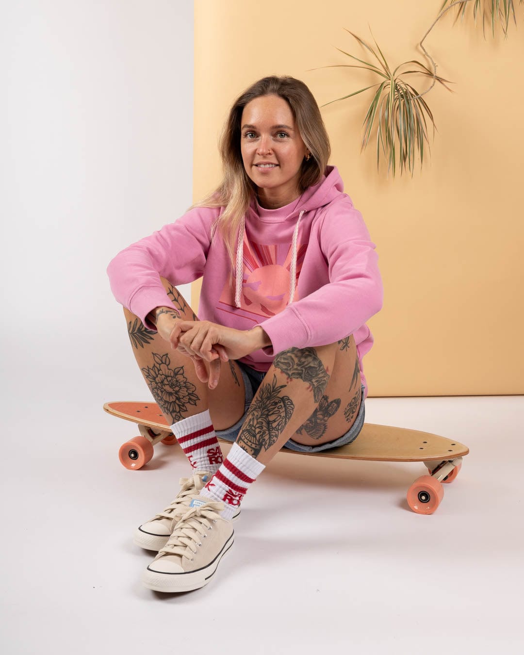 A woman wears the Saltrock Open Spaces Women's Pop Hoodie in pink while sitting on a skateboard. She has floral leg tattoos, striped socks, and sneakers. A plant and beige backdrop are behind her.