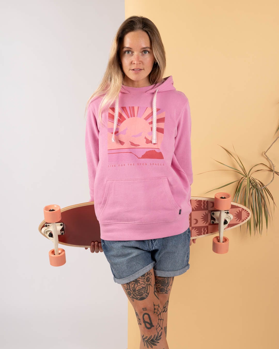 A woman in the Saltrock Open Spaces Women's Pop Hoodie in pink and denim shorts holds a skateboard against a beige and yellow backdrop with a potted plant, reflecting the easygoing Saltrock Sun vibe.