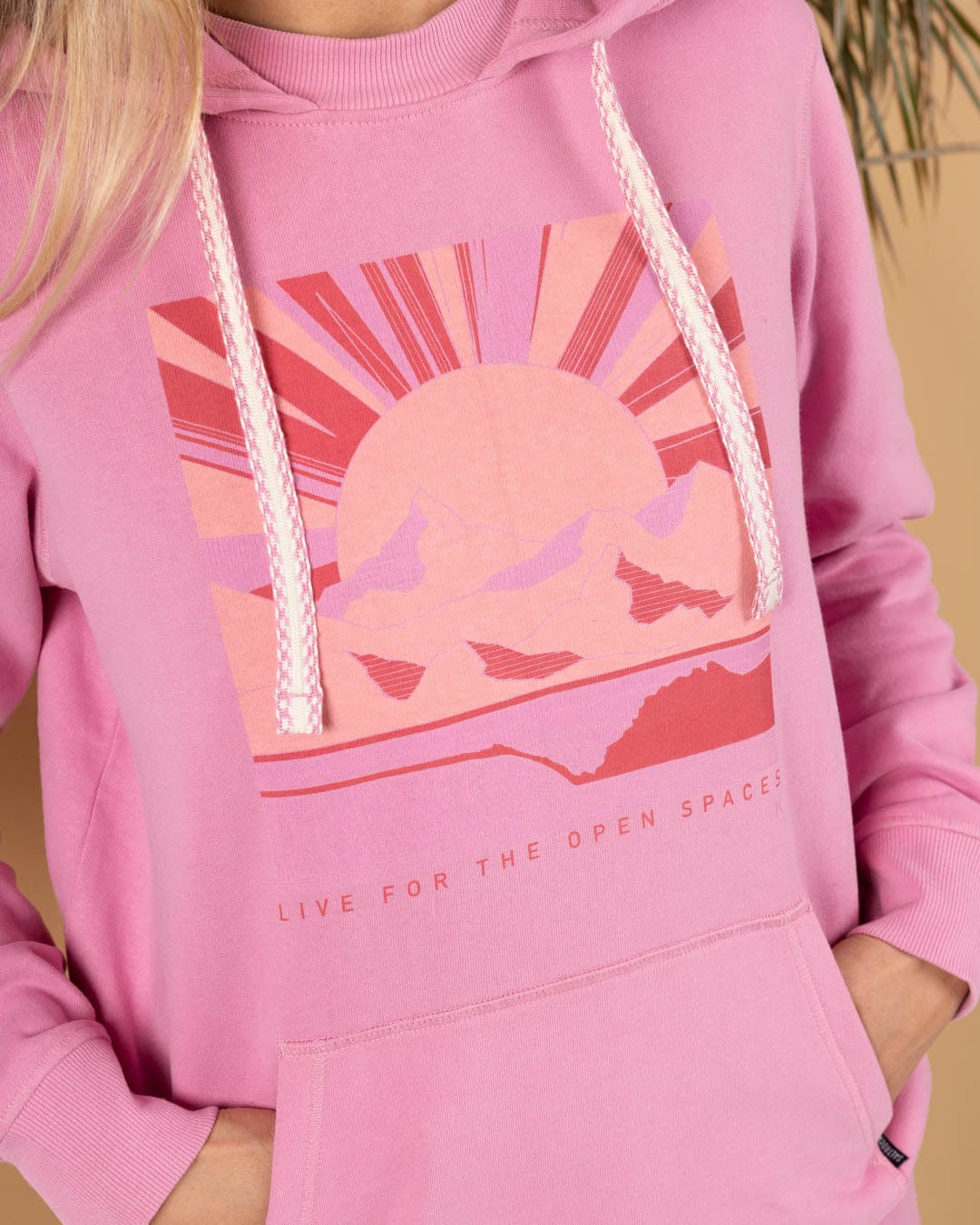 A person wears the Saltrock Open Spaces Women's Pop Hoodie in pink, featuring a sun and mountains graphic with the text "LIVE FOR THE OPEN SPACES" on the front.