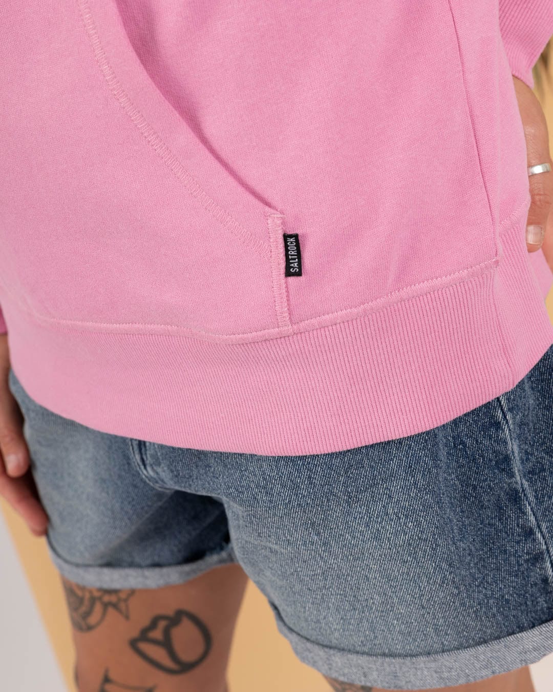 A person wears the Saltrock Open Spaces - Womens Pop Hoodie in pink with blue denim shorts, revealing a tattooed leg and hand adorned with a silver ring.