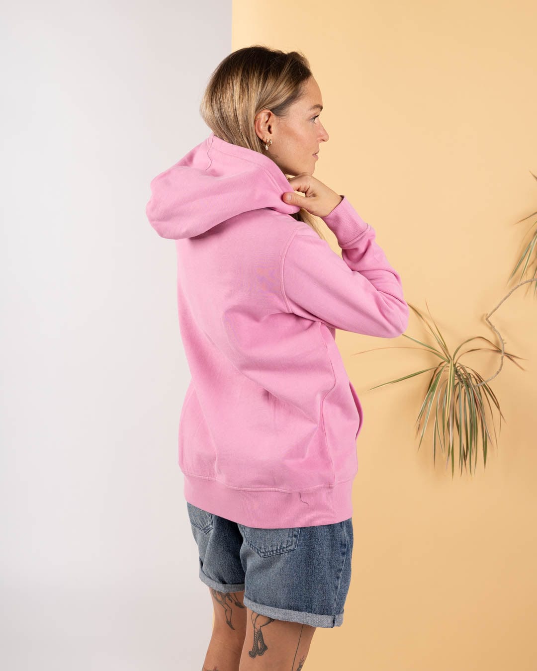 A person wearing the Saltrock Open Spaces - Womens Pop Hoodie - Pink and denim shorts stands sideways against a two-tone beige and white background with a plant.
