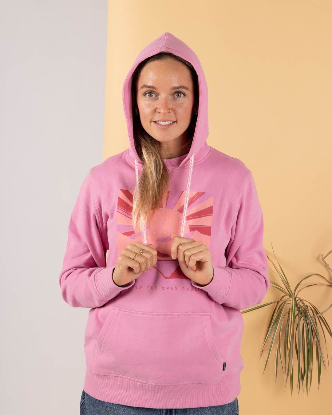 A woman stands indoors against a two-tone background, wearing the Saltrock Open Spaces - Womens Pop Hoodie in pink and holding the hoodie strings. A plant is partially visible on the right.