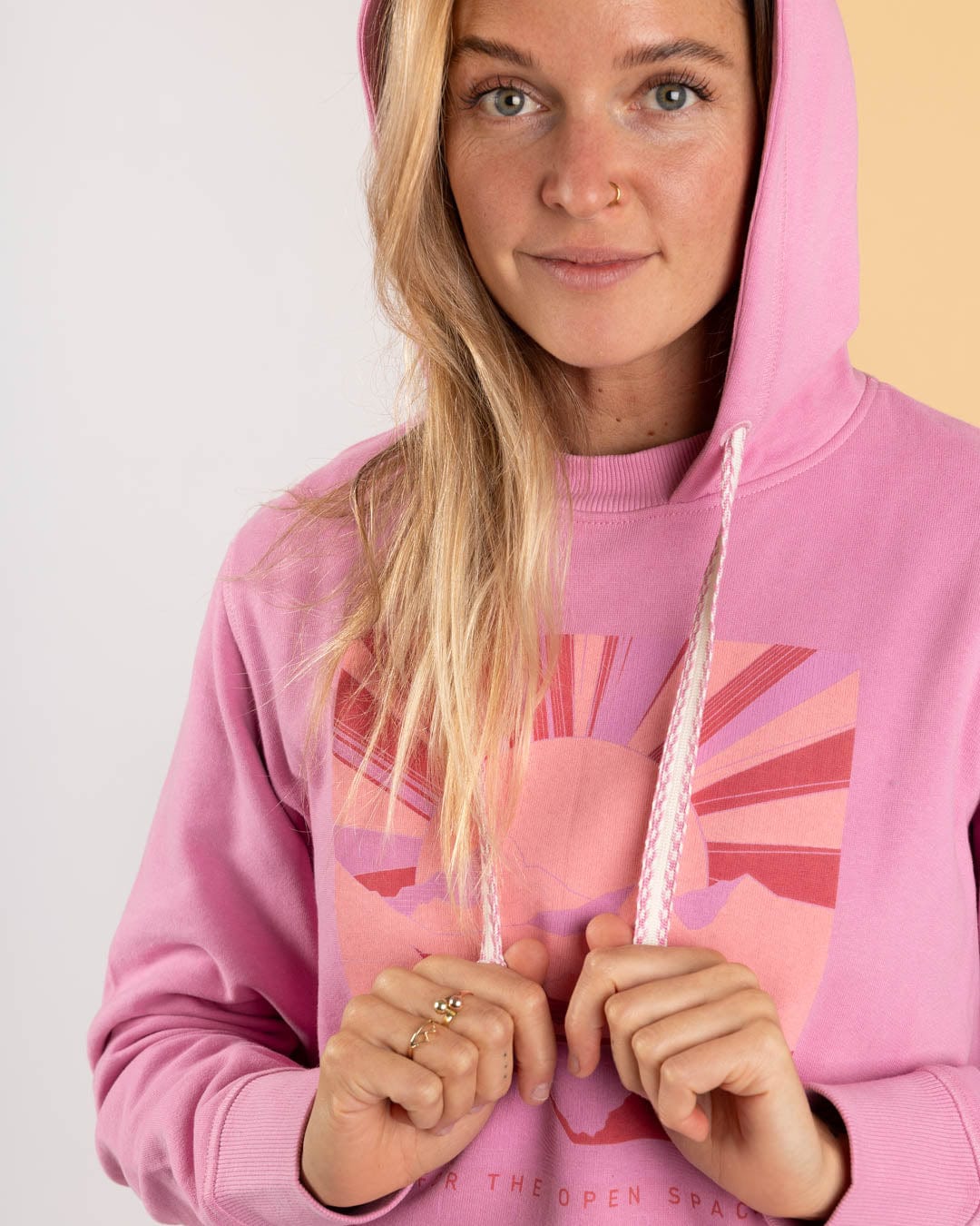 A woman wearing the Saltrock Open Spaces - Womens Pop Hoodie in pink holds the strings and looks at the camera, showcasing its graphic front design.