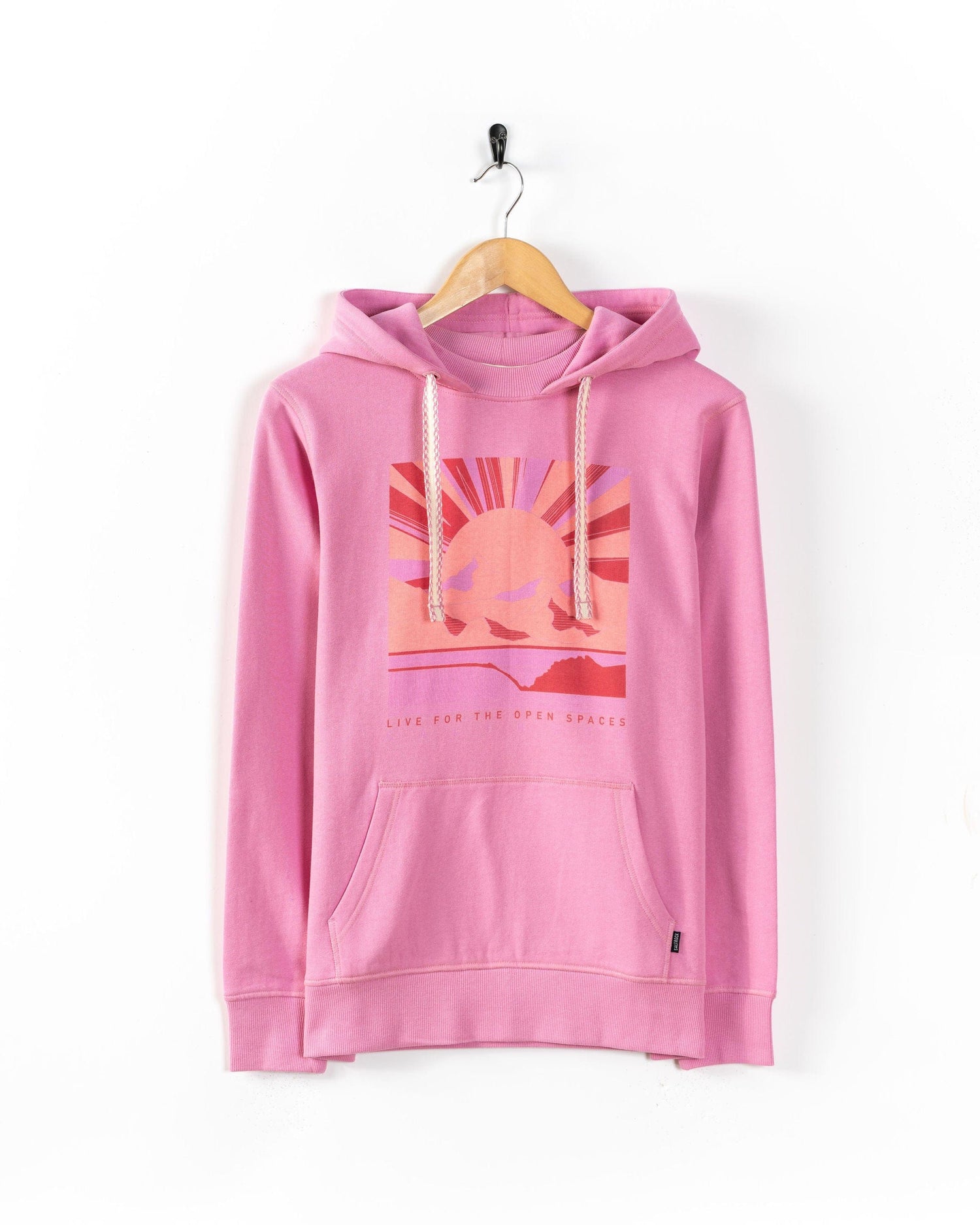 The Saltrock Open Spaces - Womens Pop Hoodie in pink features a front pocket, white drawstrings, and a Sun over mountains graphic with "Live for the open spaces," shown on a wooden hanger against a white wall.