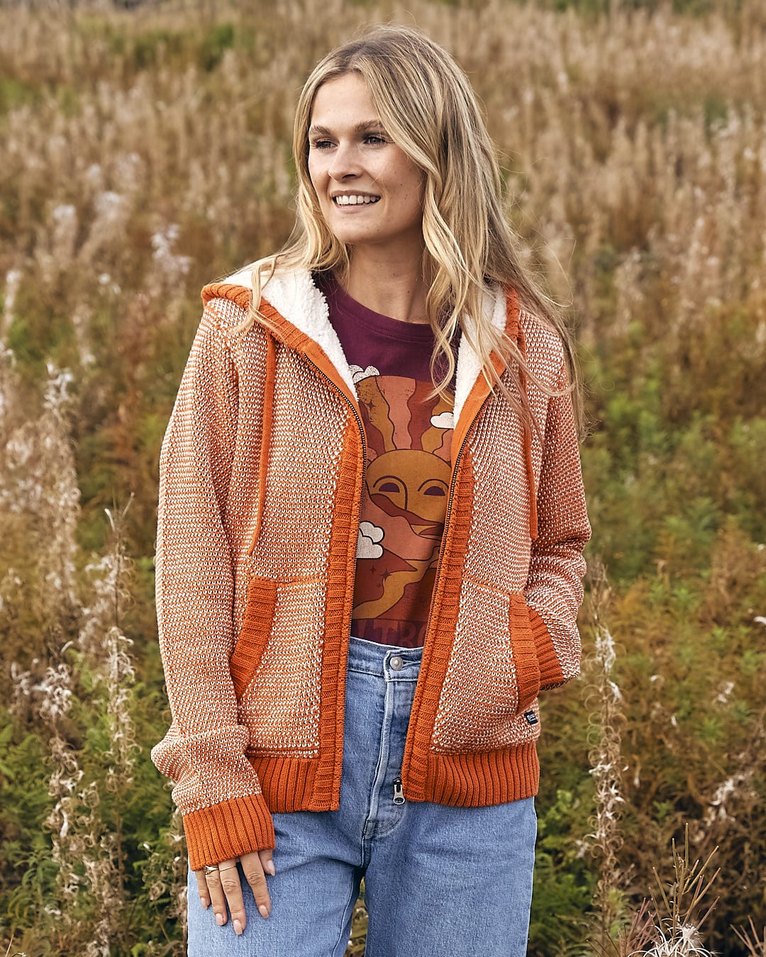 A woman in a Saltrock Helly - Womens Borg Lined Knitted Hoodie - Orange sweater and jeans standing in a field.