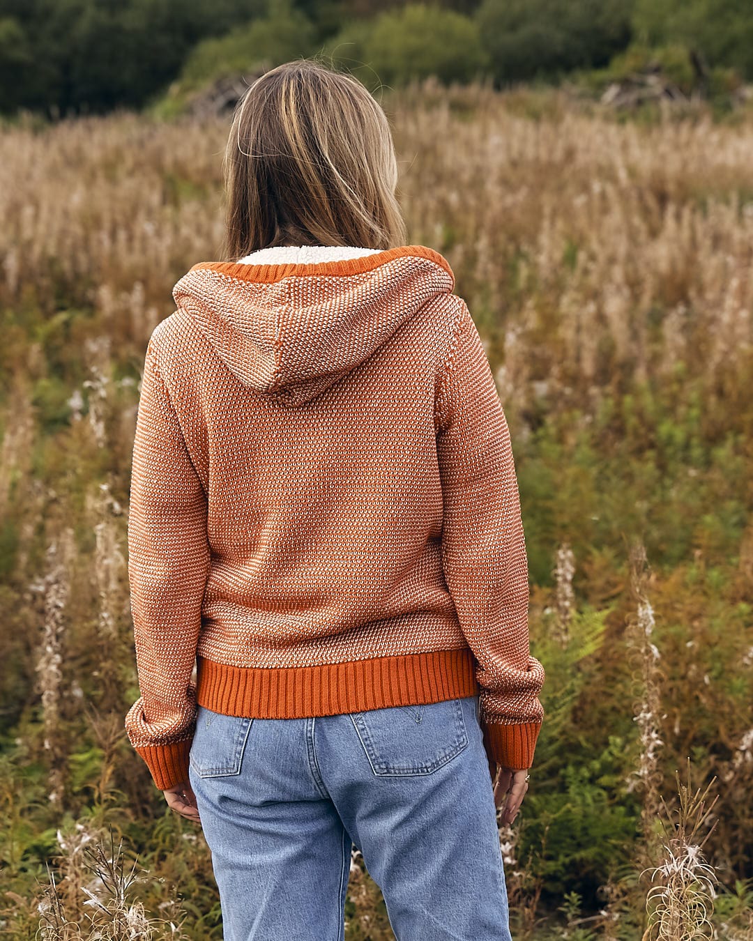 A woman standing in a field wearing a Saltrock - Helly Womens Borg Lined Knitted Hoodie - Orange sweater and jeans.