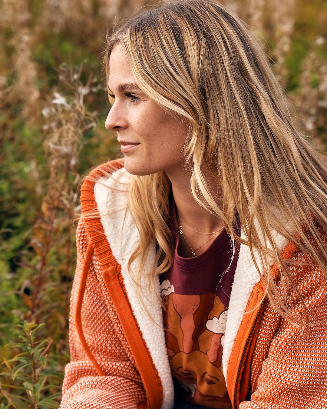 A woman sitting in a field with long blonde hair is wearing the Saltrock - Helly Womens Borg Lined Knitted Hoodie in Orange.