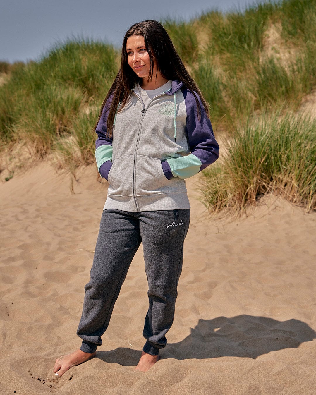 A girl standing on a sand dune in a Saltrock Jan Womens Zip Hoodie - Grey and sweatpants, exuding effortless style.