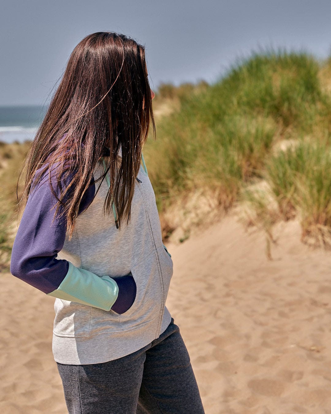 A woman standing on a sand dune effortlessly sporting a Jan - Womens Zip Hoodie - Grey, featuring the iconic Saltrock branding.