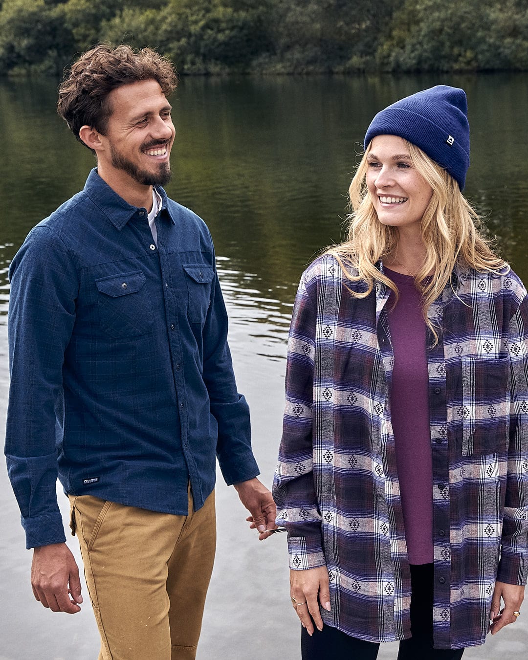 A man and woman standing next to a body of water wearing the Saltrock Jaxon - Mens Check Long Sleeve Shirt in Dark Blue.