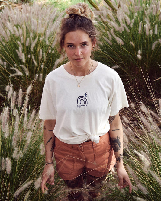 A woman with tattoos stands in tall grass, wearing a relaxed fit white Saltrock Journey - Recycled Womens Short Sleeve T-Shirt - Cream with a knotted front and embroidered graphics, along with light brown shorts made from recycled material. She has her hair up in a bun and is looking directly at the camera.