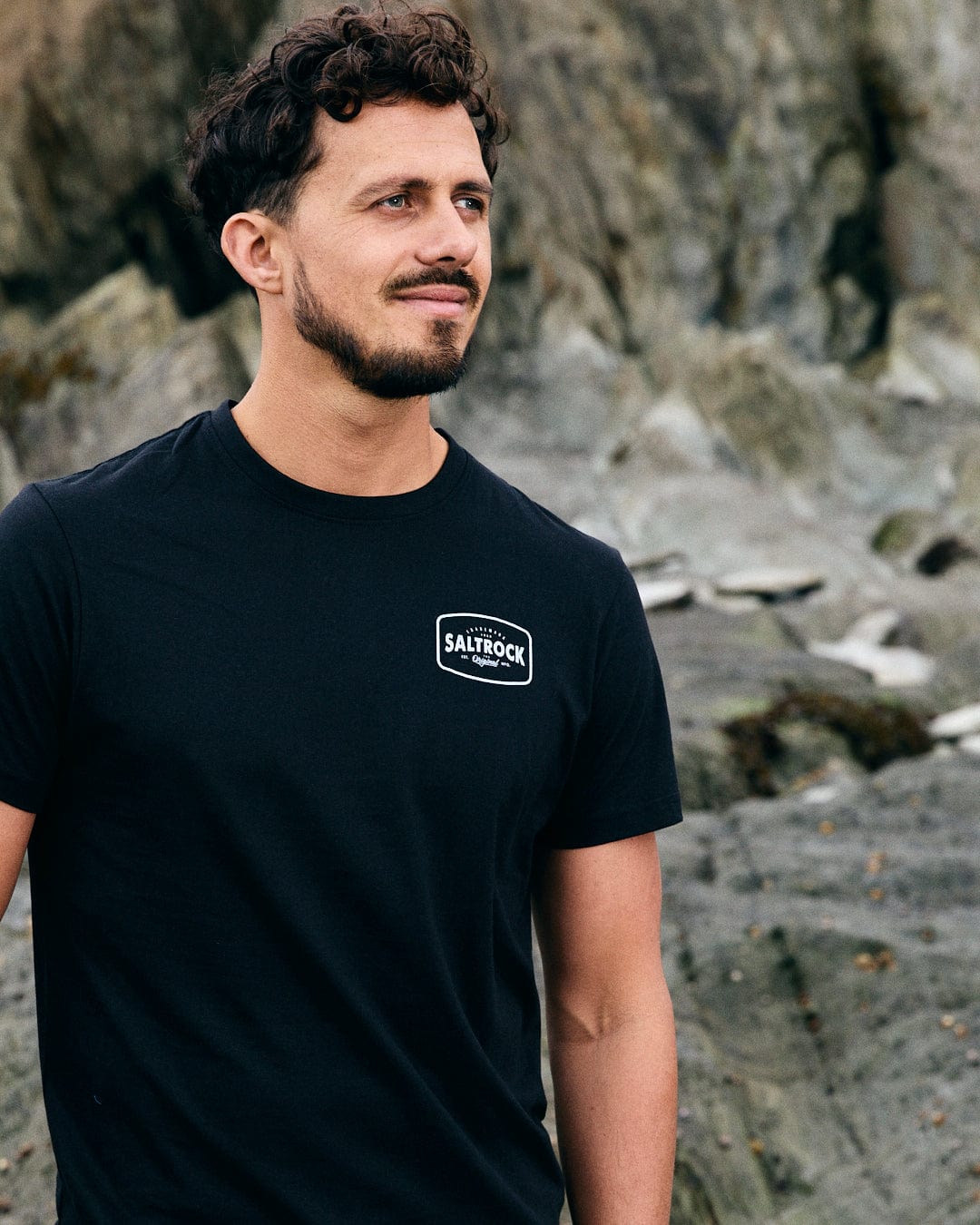 A bearded man stands on a rocky beach in a black Saltrock Jungle Wilds 2 t-shirt made from 100% cotton, gazing into the distance.