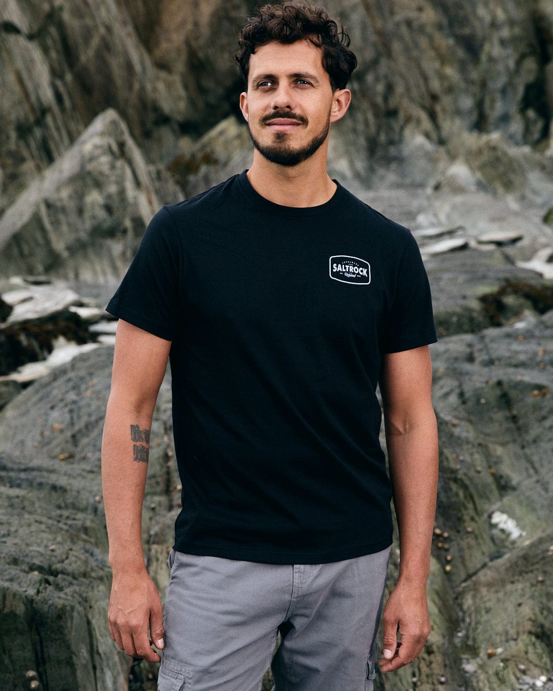 A bearded man with curly hair stands on rocky terrain, wearing the Saltrock Jungle Wilds 2 men's black T-shirt and gray shorts, showcasing his adventurous spirit.