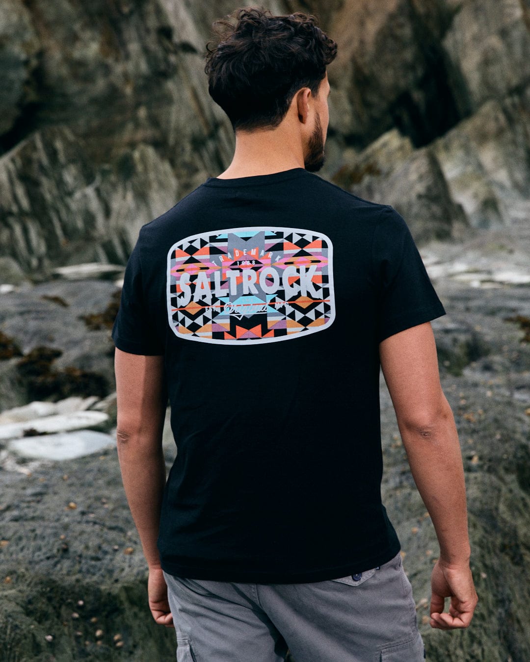 A man stands on a rocky beach wearing a Saltrock Jungle Wilds 2 T-shirt in black, featuring an Aztec-inspired geometric design on the back, made from 100% cotton.