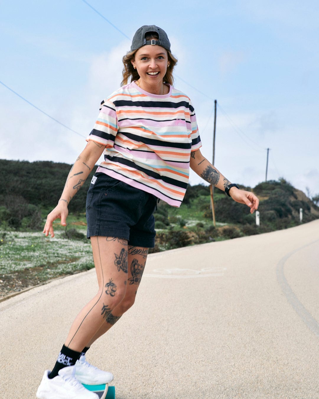 A young woman in a Saltrock dark grey striped Palm Cap and shorts skating on a road, smiling, with tattoos visible on her legs and arms.