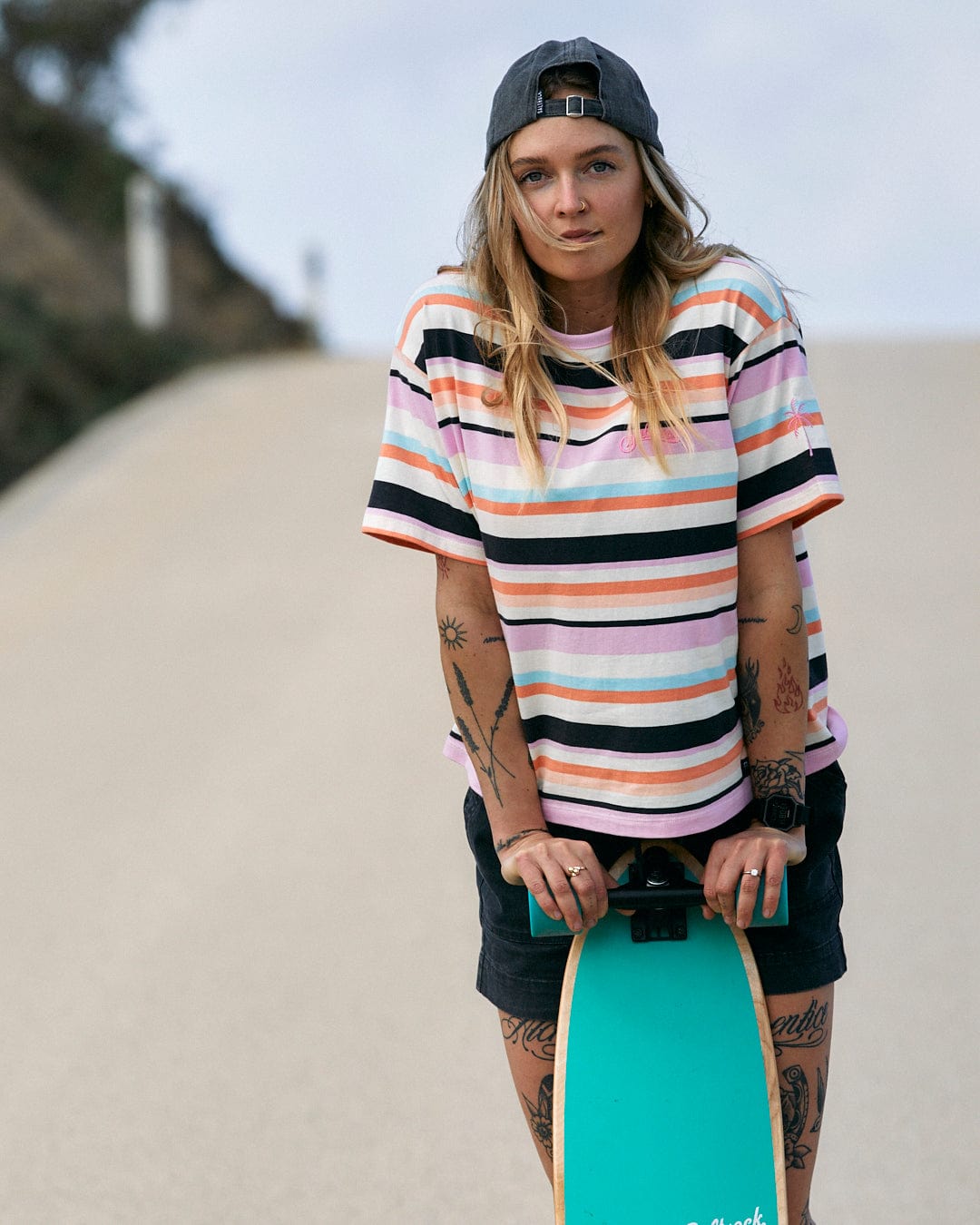 A person holding a skateboard on a paved path, wearing a Saltrock Juno - Womens Short Sleeve T-Shirt - Multi with a stripe pattern and a baseball cap.