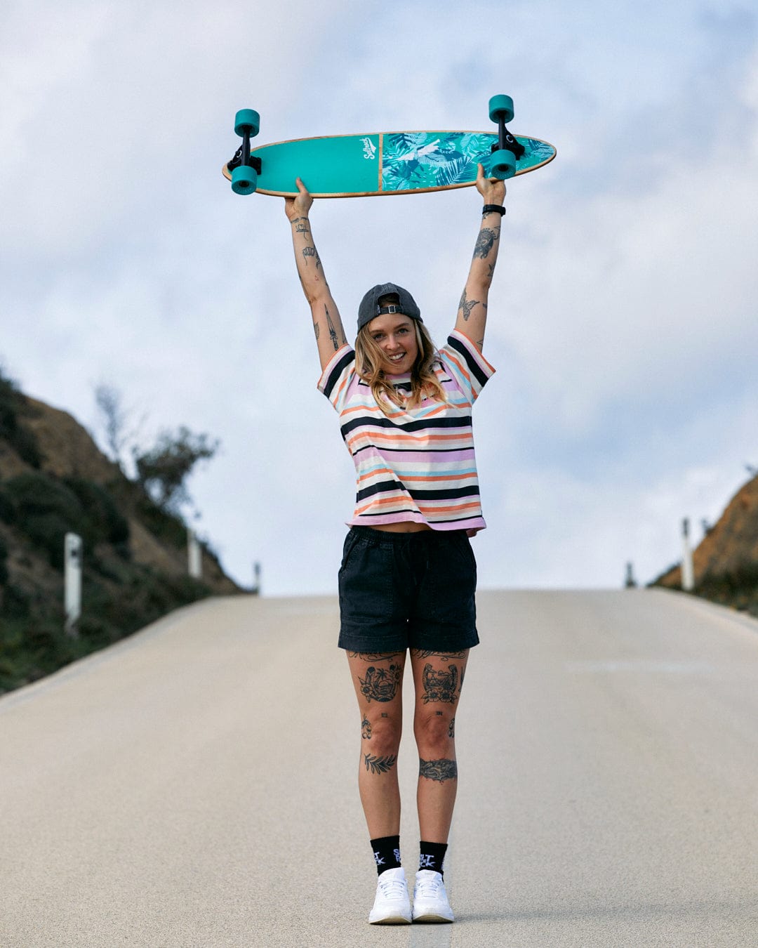 A person holding a skateboard above their head on a road, wearing a Saltrock Juno - Womens Short Sleeve T-Shirt - Multi with an embroidery stripe pattern.