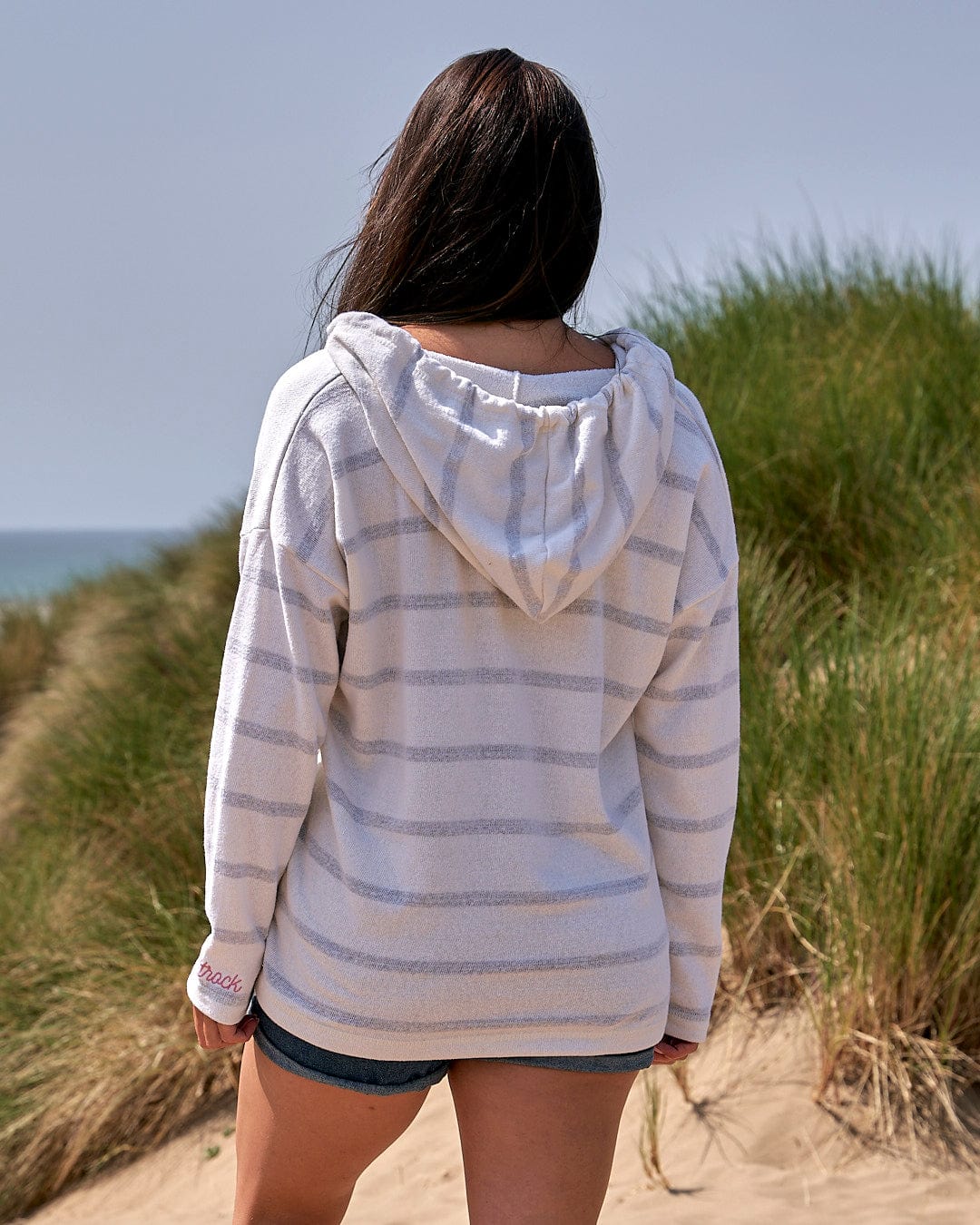 A woman wearing a Saltrock Kennedy - Womens Pop Hoodie - Cream, with white stripes, walking on a sand dune.