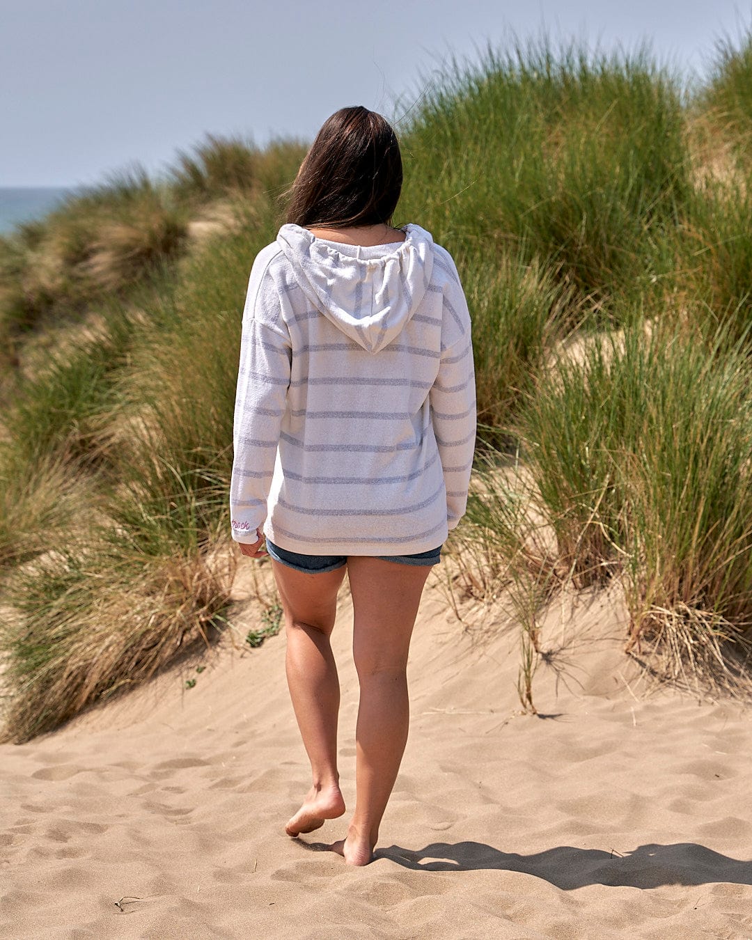 A woman walking on a sand dune near the ocean wearing the Saltrock Kennedy - Womens Pop Hoodie - Cream.