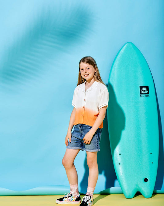 A young girl in a Saltrock Raya - Kids Shirt - Orange, denim shorts, and sneakers stands beside a turquoise surfboard against a blue backdrop.