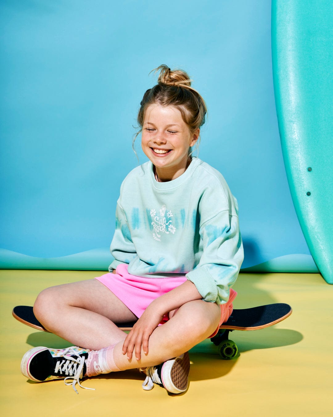 A smiling child with a top bun hairstyle sits cross-legged on a skateboard, wearing a Peace Love - Kids Sweat - Green sweatshirt by Saltrock, pink shorts, and black sneakers. In the background, a teal surfboard stands tall alongside a tie-dye stripe that reads "Peace Love Saltrock.