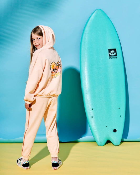 A girl in the Sunshine State - Kids Zip Hoodie - Peach with matching pants stands next to an aqua blue Saltrock branded surfboard in a studio with a blue background.