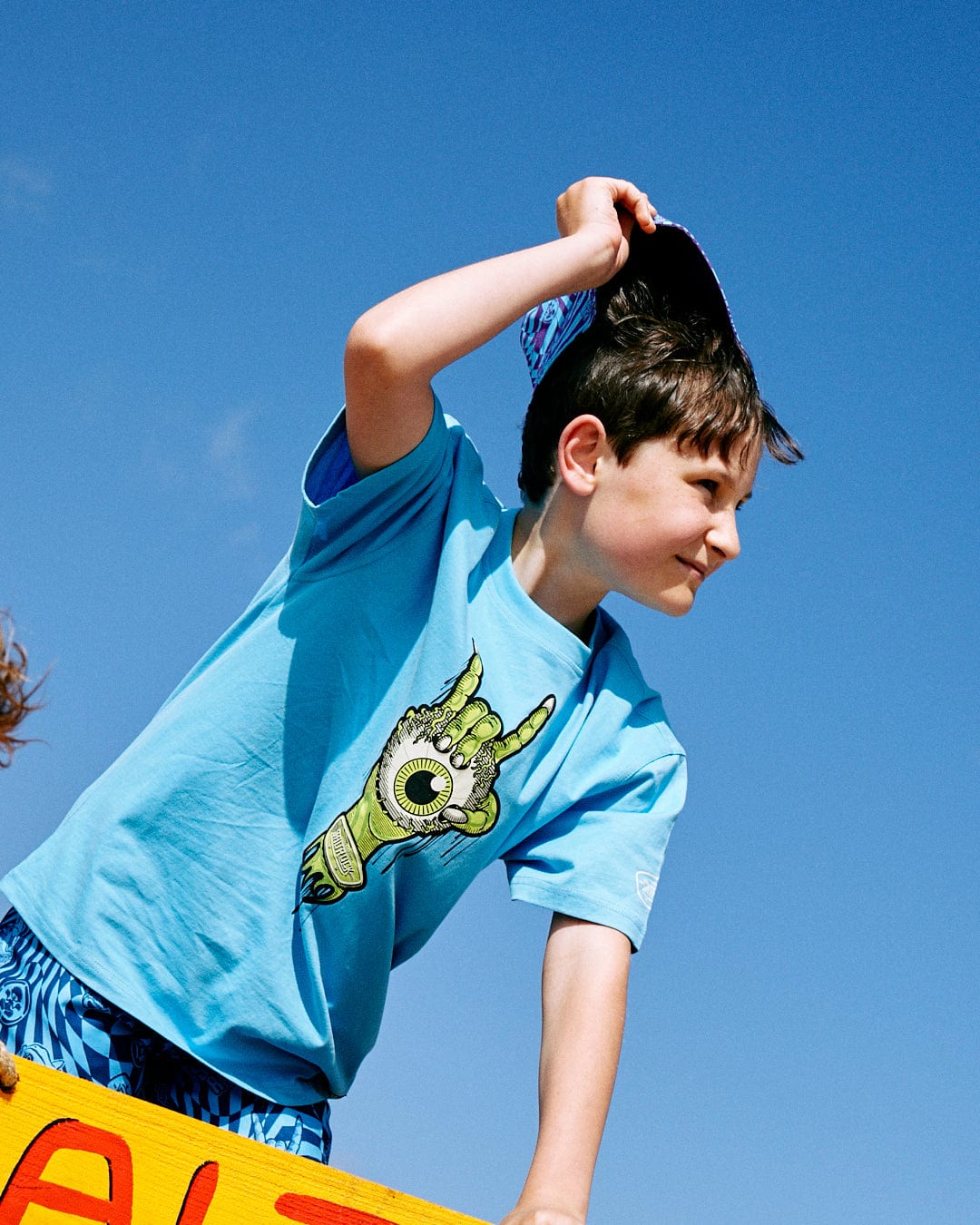 A young boy in a 100% cotton blue Bowling for Surf - Kids Short Sleeve Glow in the Dark T-Shirt from Saltrock with a graphic of a hand on it adjusts his blue cap under a clear blue sky.