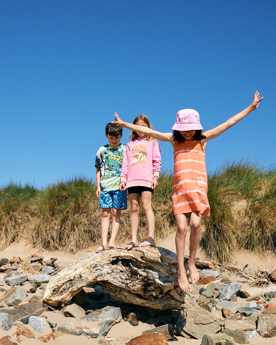 Three children stand on a driftwood log at the beach. One child in an Eliana - Kids Tie Dye Vest Dress - Peach by Saltrock and hat has arms outstretched, while the other two stand behind, dressed in casual summer outfits.