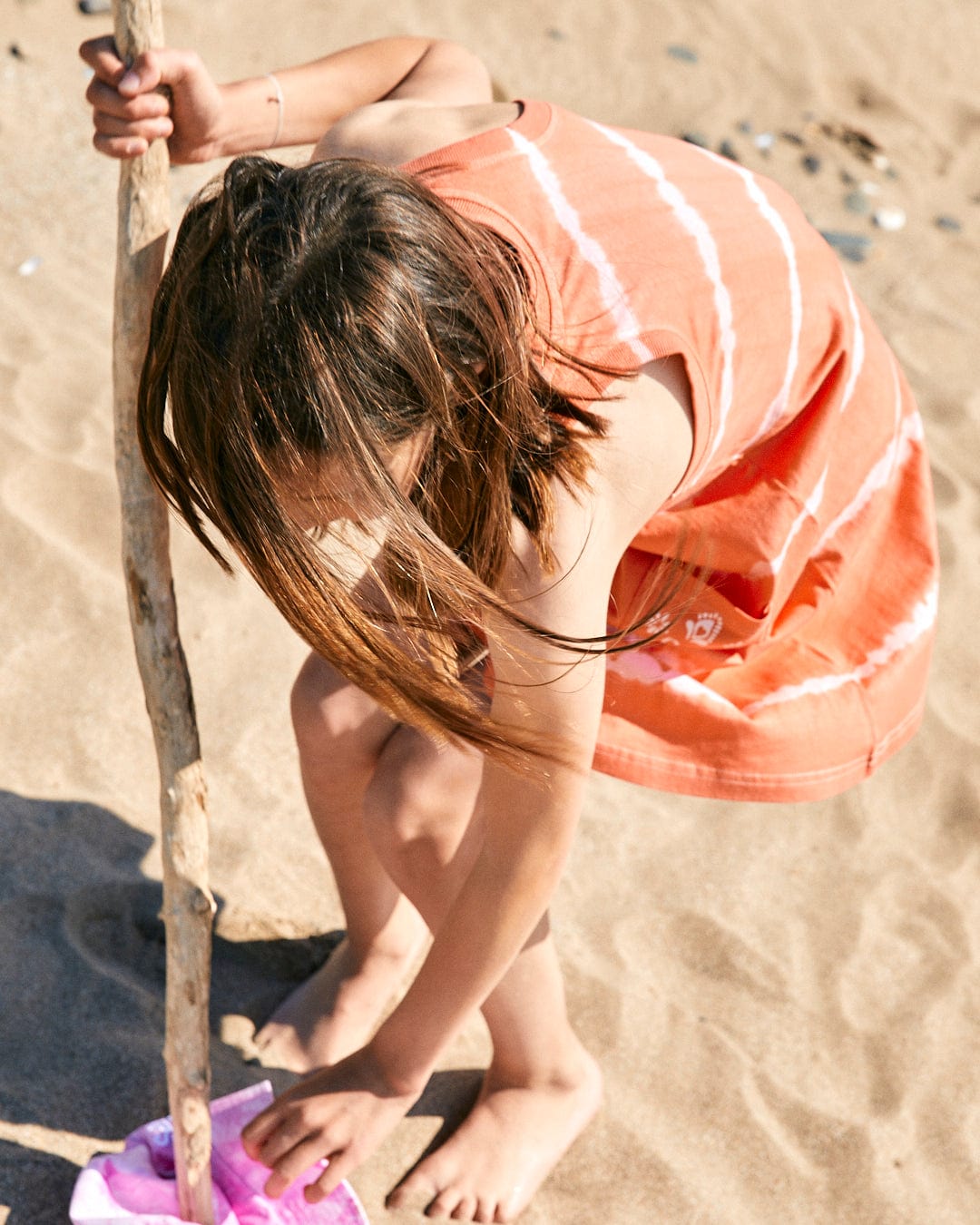 A child in a Saltrock Eliana - Kids Tie Dye Vest Dress - Peach is bending down to play in the sand while holding a stick on a sunny beach.