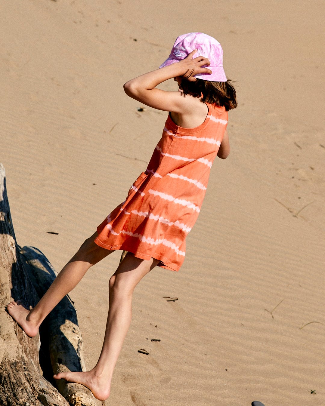 A child wearing the Saltrock Eliana - Kids Tie Dye Vest Dress - Peach and a purple hat walks barefoot on the sand next to a log.