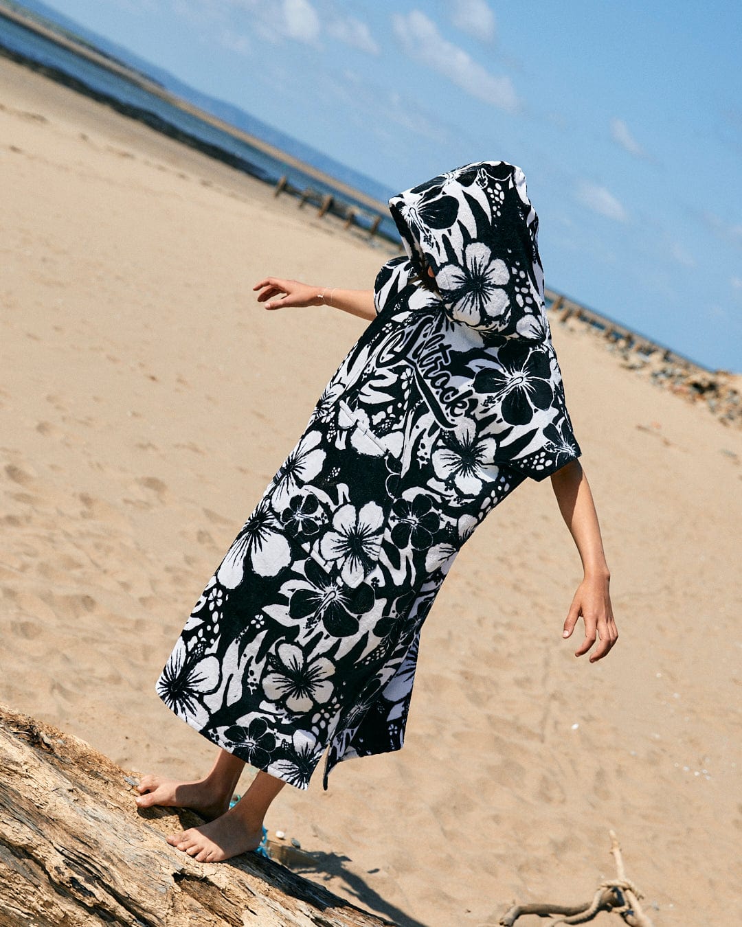 A child on a sandy beach wearing a Saltrock Hibiscus - Kids Changing Towel - Black/White, 100% cotton and ultra absorbent, stands near a piece of driftwood with the blue sky and distant water in the background.