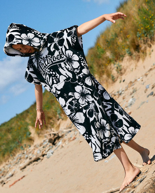 A child wearing a Saltrock Hibiscus - Kids Changing Towel - Black/White walks barefoot on a sandy beach with greenery in the background. The 100% cotton material is ultra absorbent, perfect for keeping them dry and comfortable.