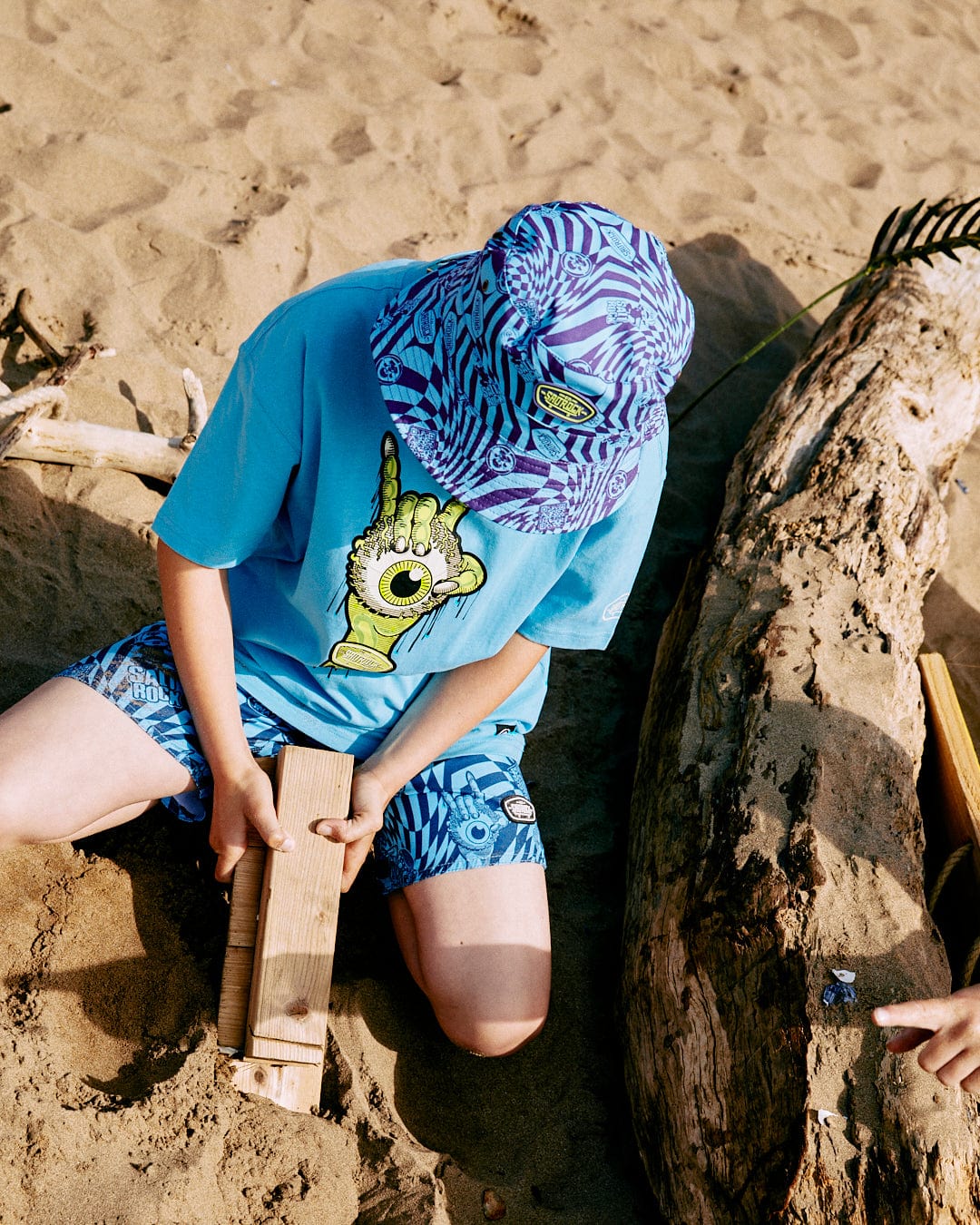 A child wearing a Bowling for Surf - Kids Short Sleeve Glow in the Dark T-Shirt by Saltrock is playing with a wooden plank on a sandy beach near a large piece of driftwood.