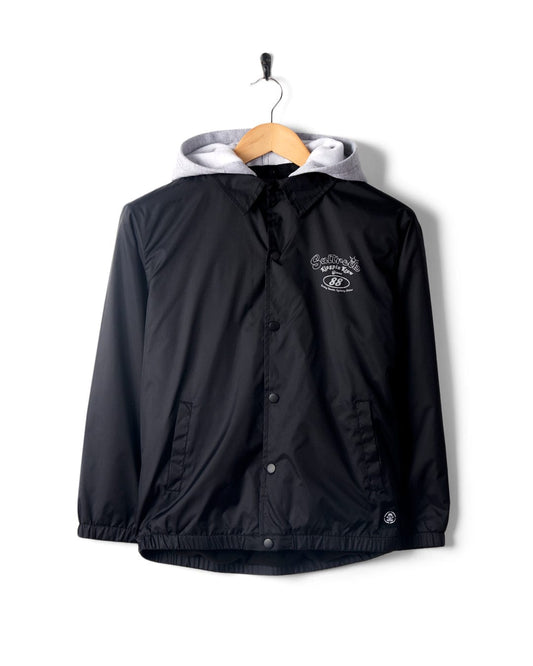 A black Kingpin Krew - Kids Coach Jacket with white detailing on the collar and a detachable hood, hanging on a wooden hanger against a white background. (Saltrock)