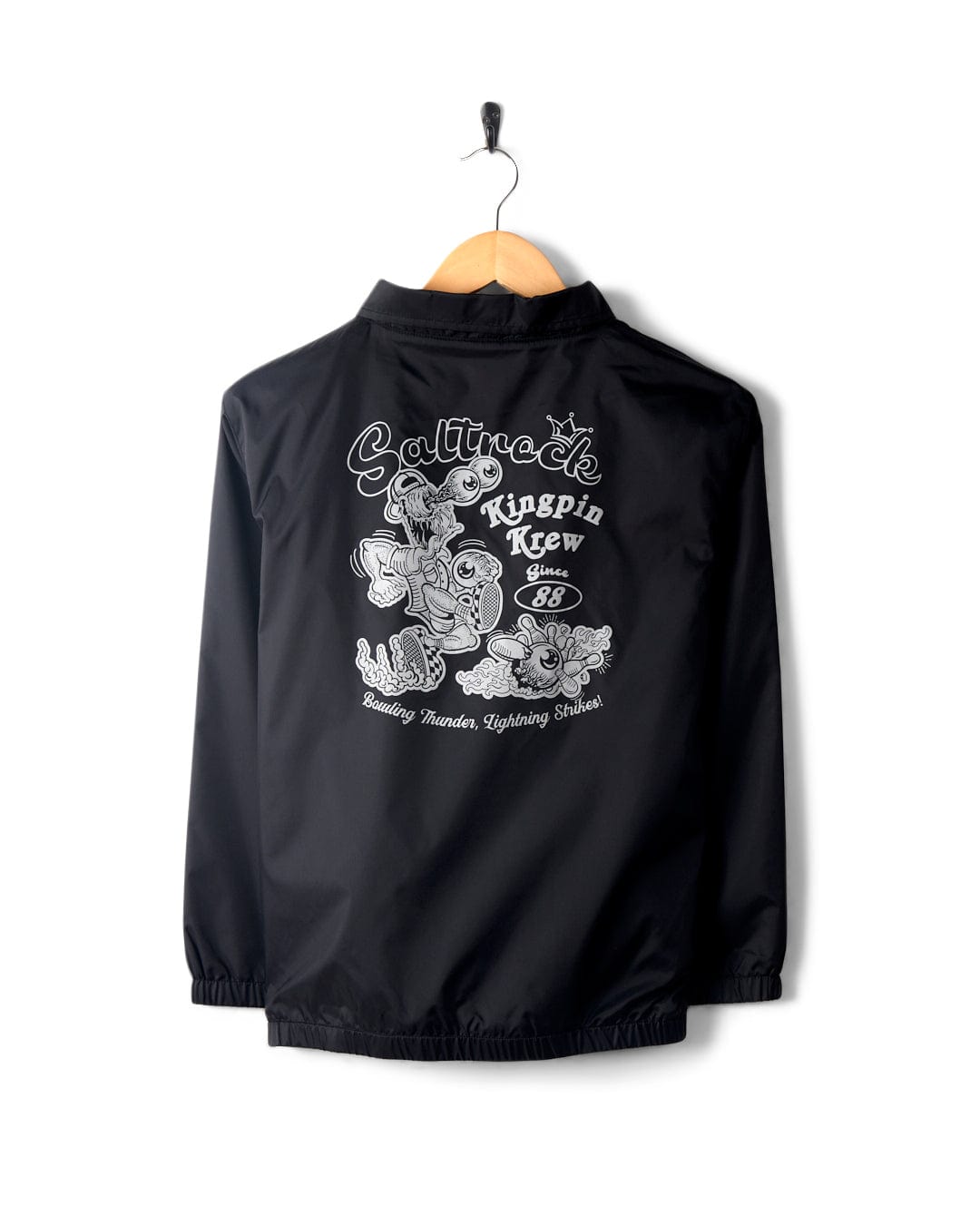 A Kingpin Krew - Kids Coach Jacket - Black with a detachable hood on a hanger featuring white graphic print with cartoon-style characters and text, displayed against a white background by Saltrock.