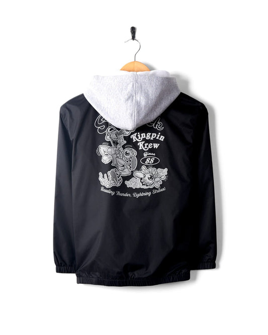 A black, water-resistant Kingpin Krew - Kids Coach Jacket - Black with graphic designs, hung on a hanger with a white Saltrock hoodie underneath, displayed against a white background.