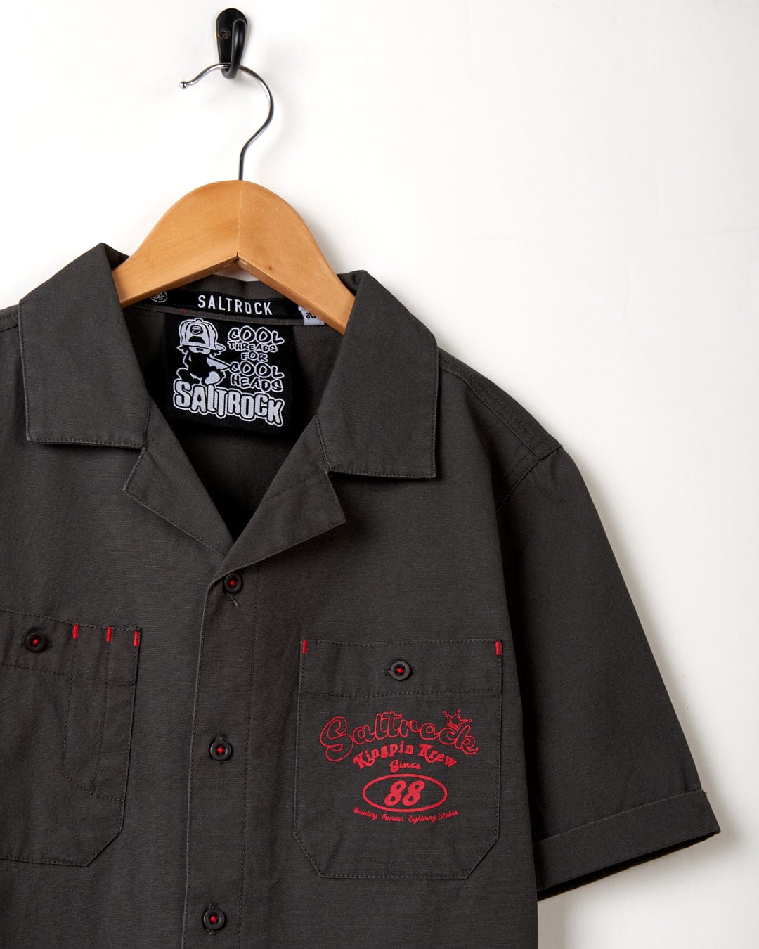 A dark grey Saltrock Kingpin Krew - Kids Short Sleeve Shirt with a red logo hanging on a hanger.