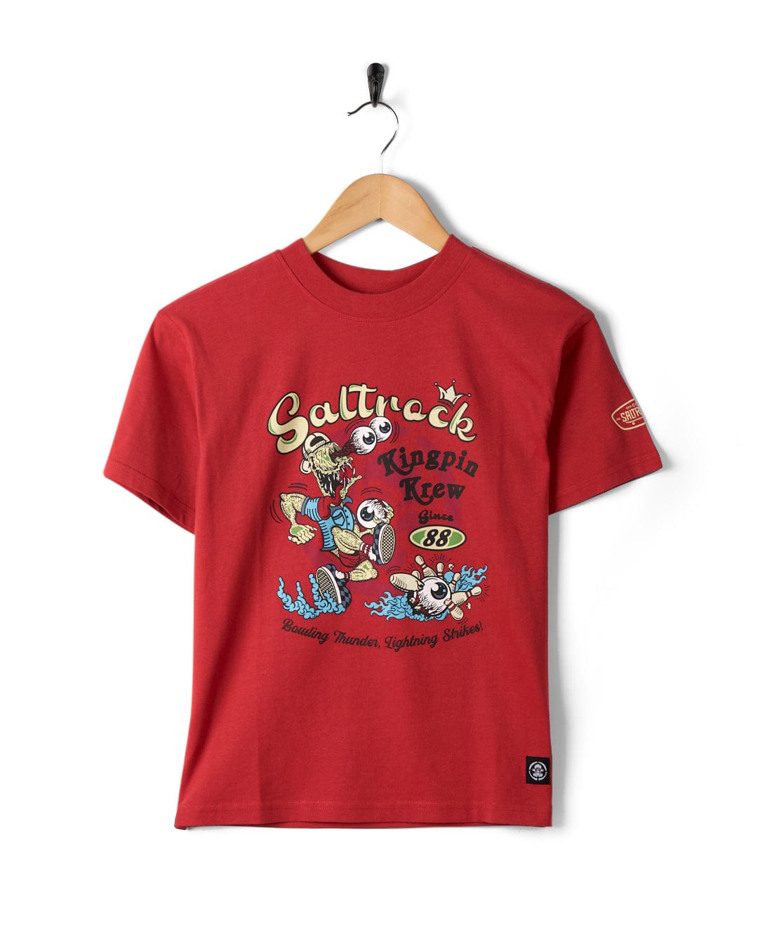 A Saltrock Kingpin Krew - Kids Short Sleeve Glow in the Dark T-Shirt on a hanger featuring a playful cartoon design with text reading "Saltrock Kingpin Krew Since '88" and "Bombing Headen, Lightning Strikes!" This relaxed fit tee is ideal for casual days.