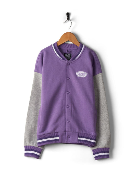 A Saltrock Krew - Recycled Kids Varsity Bomber Jacket - Purple in purple and gray with striped cuffs and collar, made from recycled material, hanging on a wooden hanger against a white background.