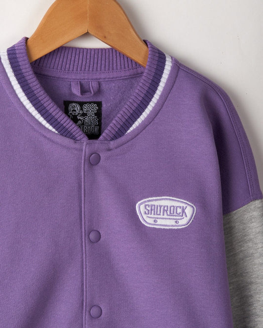Purple and grey lightweight Krew - Recycled Kids Varsity Bomber Jacket - Purple with white detailing, branded "Saltrock" on the chest. Made from recycled materials, this stylish piece also features a Saltrock skateboard applique. The jacket is hanging on a wooden hanger.