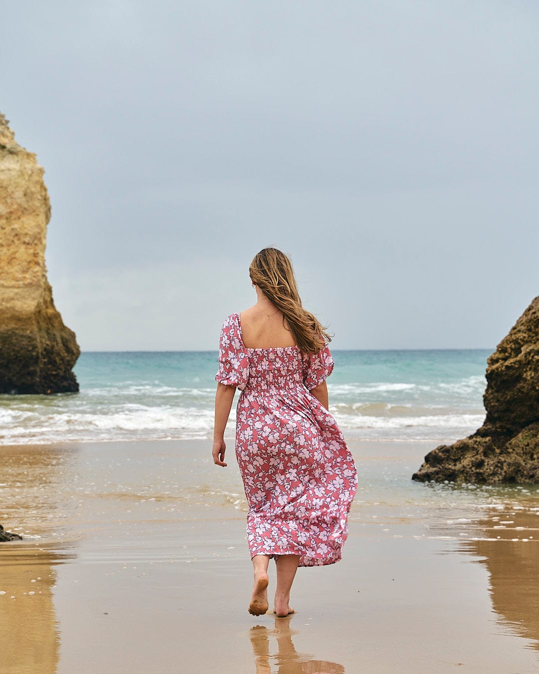 A woman in a Larran - Womens Shirred Dress - Mid Pink by Saltrock walking on the beach.
