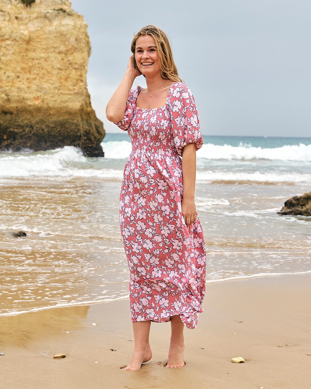 A woman in a Saltrock Larran - Womens Shirred Dress - Mid Pink standing on the beach.